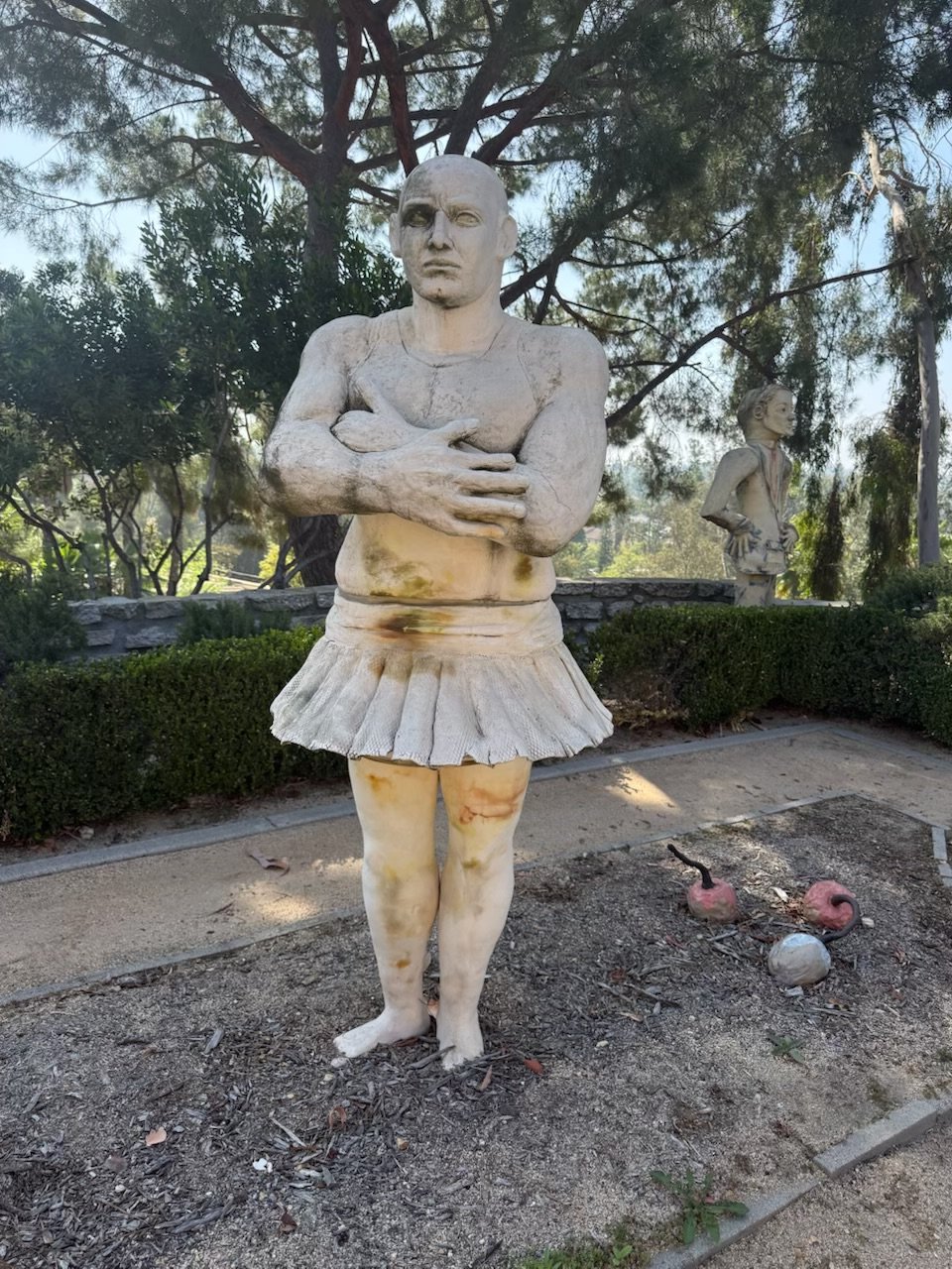 Life-size ceramic sculpture of a muscular figure in a tulle skirt, arms crossed, installed in a garden