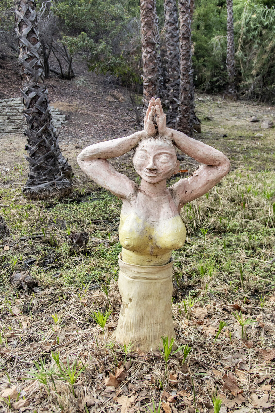 Ceramic figure with hands raised to its head standing among tall palm trunks in golden light