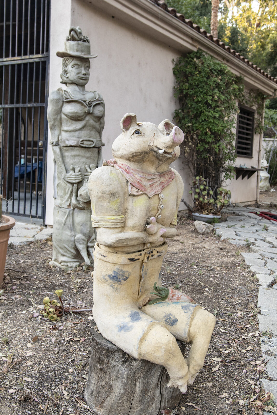 Ceramic pig in a red bandana beside a seated companion figure in a hat near a courtyard wall