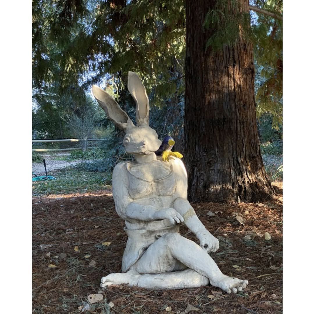 Seated white ceramic rabbit figure beneath a redwood, with a live bird perched on its shoulder