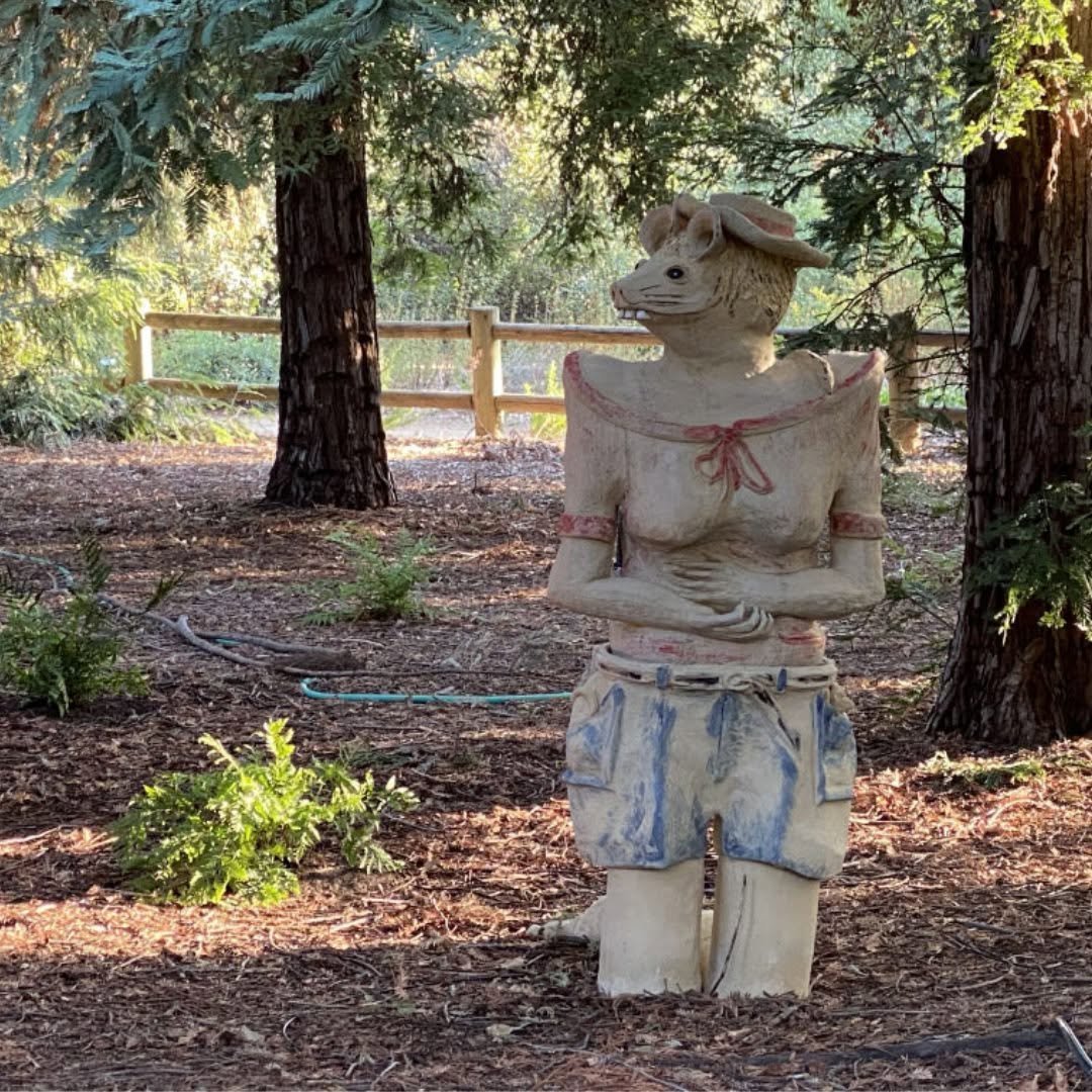 Ceramic figure with a rodent head wearing a hat, blouse and blue shorts standing among redwoods